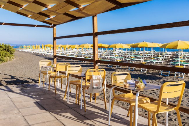 Tavoli e sedie gialle all'aperto, ombrelloni gialli sulla spiaggia in lontananza.