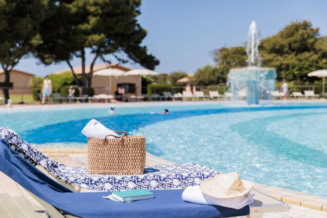 Lettino da piscina con borsa, cappello, e libro, a bordo vasca. Sullo sfondo, fontana e ombrelloni.