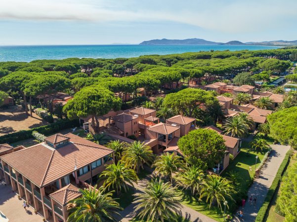 Resort con edifici dal tetto rosso circondato da pini e palme, vicino al mare.