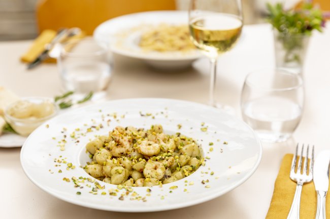 Piatto di gnocchi con gamberi e pistacchi, bicchiere di vino bianco accanto.