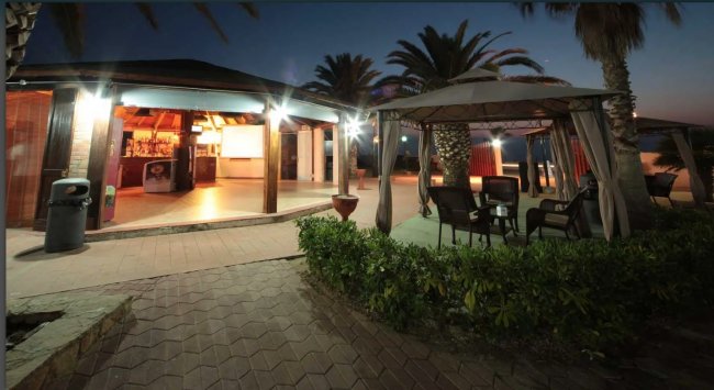 Esterno di un bar illuminato di sera con gazebo e sedie sotto le palme.
