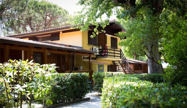 Edificio a due piani in legno e muratura con balcone, circondato da alberi e arbusti.