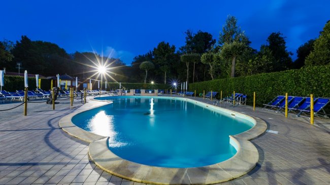 Piscina illuminata di notte con lettini blu e siepi intorno.