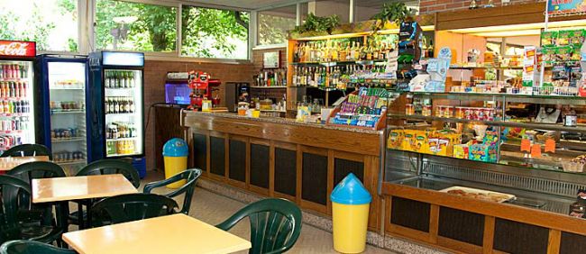 Interno di un bar con tavoli, sedie verdi e un bancone fornito di bevande e snack.