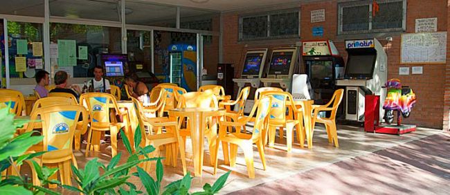 Sala giochi con tavoli gialli e sedie, videogiochi arcade al fondo.