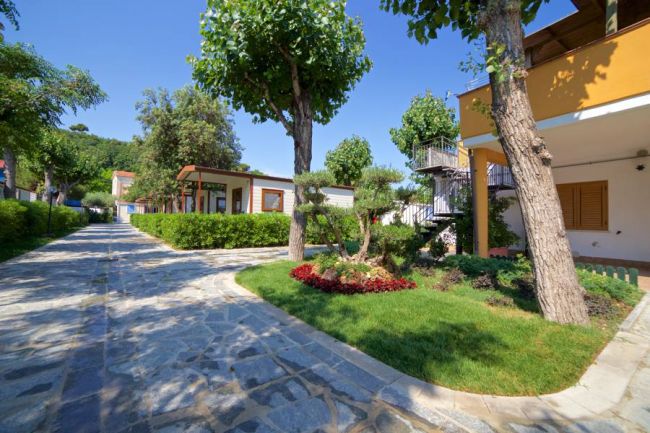 Un viale acciottolato tra giardini curati e bungalow colorati, circondati da alberi.