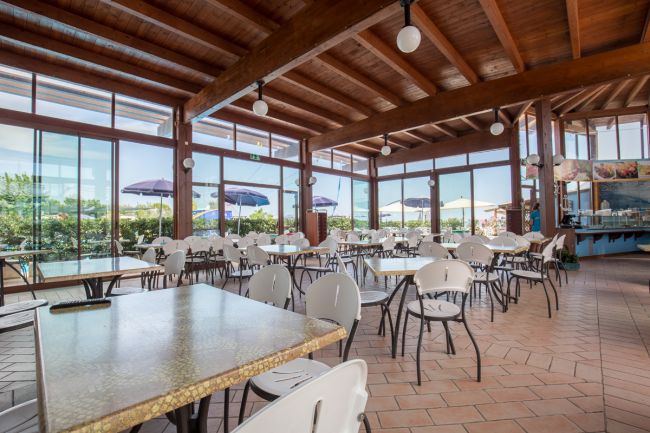 Una sala da pranzo con tavoli e sedie bianche, soffitto in legno e ampie finestre.