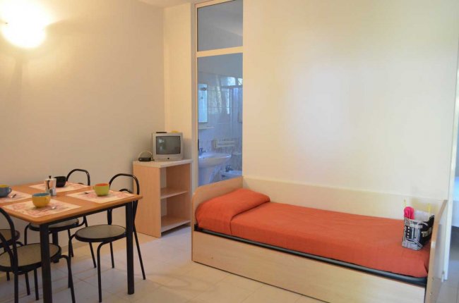 Stanza con letto singolo arancione, tavolo da pranzo nero e piccola TV su mobile.