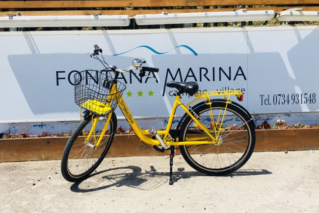 Una bicicletta gialla con cestino nero parcheggiata davanti a un cartello.