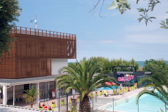 Una piscina esterna con palme, un edificio moderno a sinistra e un cartello di benvenuto colorato.