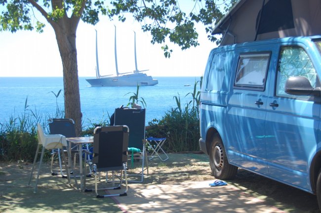 Un furgone blu accanto al mare con un'imbarcazione a vela in lontananza.
