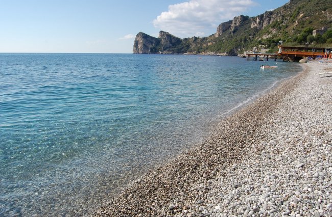 Spiaggia di ciottoli con mare limpido e scogliere sullo sfondo.