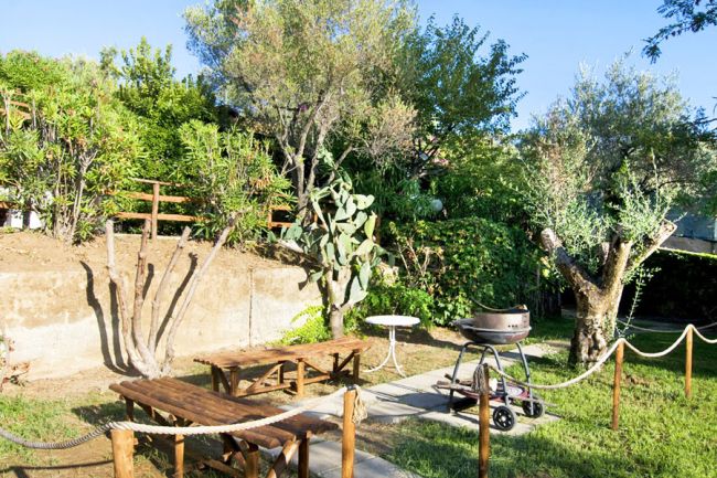 Un giardino con barbecue, tavoli in legno, vegetazione rigogliosa e un sentiero recintato.