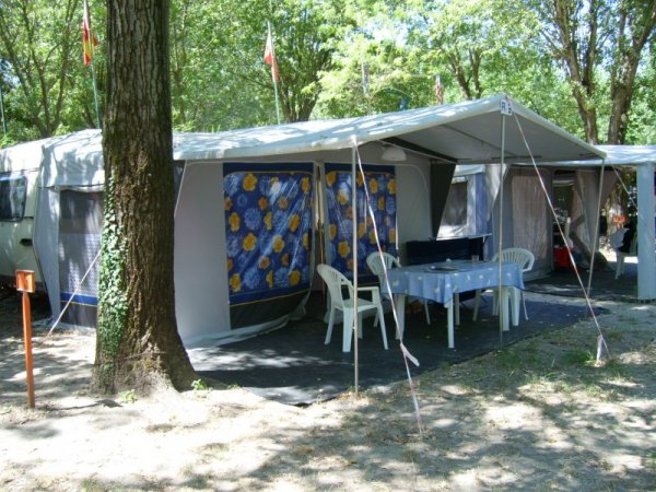 Una tenda campeggio con tavolo e sedie bianche sotto un baldacchino, circondata da alberi.