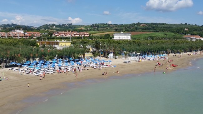 Spiaggia con ombrelloni blu, bagnanti, mare calmo, colline verdi sullo sfondo.