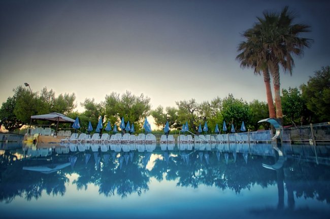 Piscina con lettini, ombrelloni blu e palme sullo sfondo.
