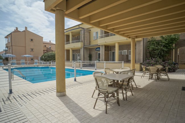 Area piscina con tavoli e sedie sotto una tettoia, edifici sullo sfondo.