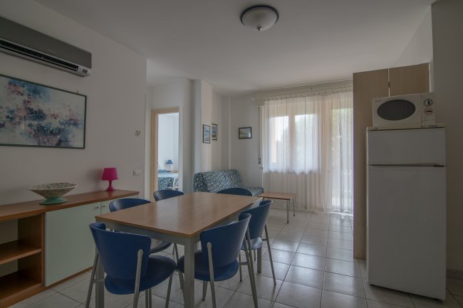 Soggiorno con tavolo e sedie blu, divano, frigorifero, quadro e aria condizionata.