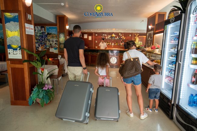 Famiglia con valigie al bancone della reception dell'hotel, vicino a un frigorifero con bevande.