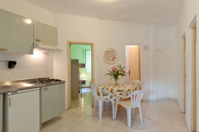 Cucina e zona pranzo con tavolo rotondo, sedie di plastica, fiori e toni chiari.