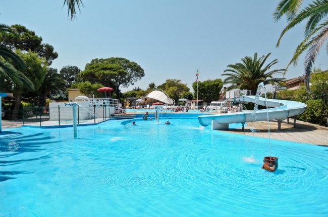 Piscina con scivolo e spruzzi d'acqua, circondata da palme e ombrelloni.