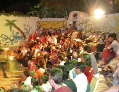 Un pubblico di persone sedute all'aperto, alcune in piedi, con un murale di sfondo.