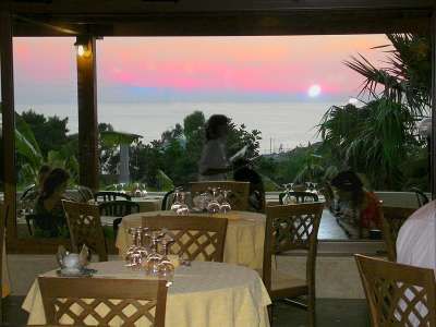 Ristorante all'aperto con tavoli apparecchiati, vista sul tramonto e mare.