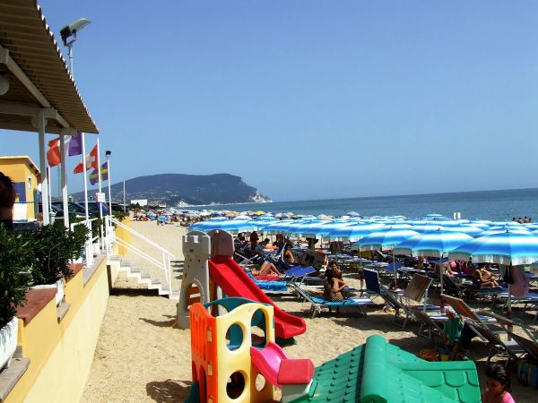 Spiaggia affollata con ombrelloni blu e giochi per bambini sullo sfondo del mare.