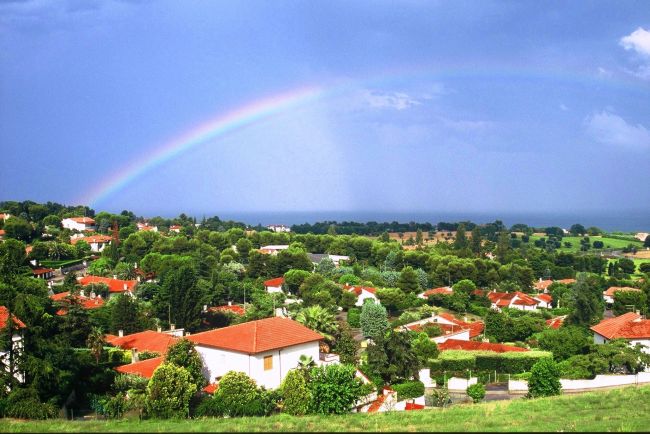 Paesaggio collinare con case dai tetti rossi, arcobaleno nel cielo.