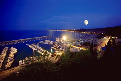 Vista notturna di un porto illuminato con una luna piena e mare calmo.