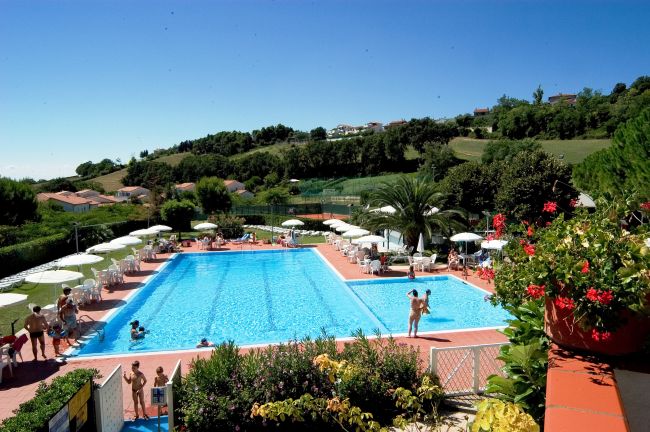 Piscina con ombrelloni, persone a bordo e colline verdi attorno.