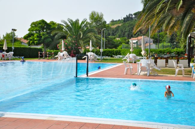Piscina all'aperto con persone che nuotano, circondata da sedie bianche e palme sullo sfondo.