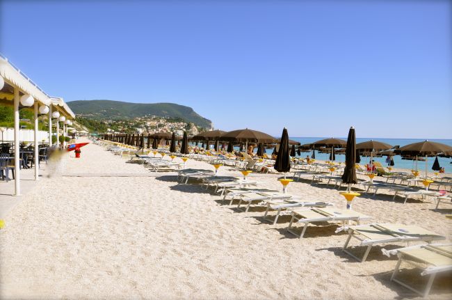Spiaggia con file di lettini e ombrelloni marroni, vista mare e colline in lontananza.