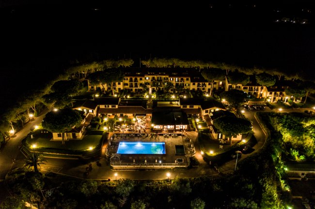 Vista aerea notturna di un complesso alberghiero con piscina illuminata e alberi attorno.