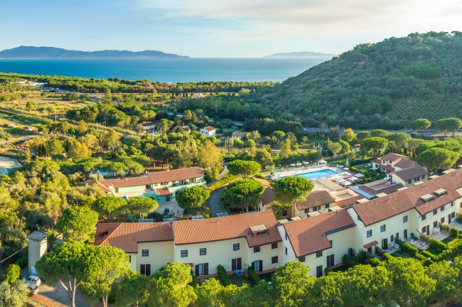 Complesso di edifici con tetti rossi circondato da vegetazione lussureggiante e vista sul mare.