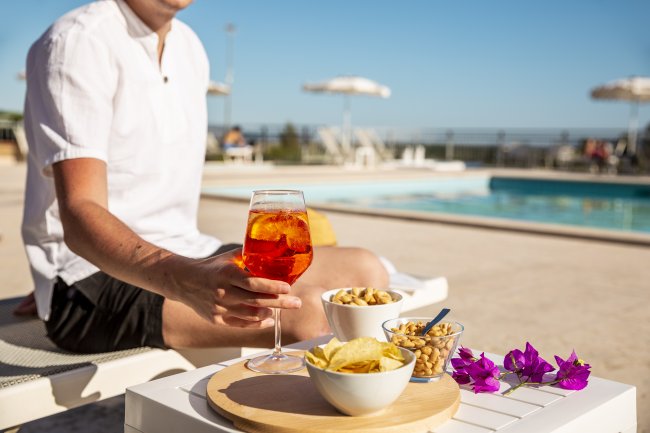 Una persona in camicia bianca tiene un drink arancione vicino a una piscina con snack e fiori.