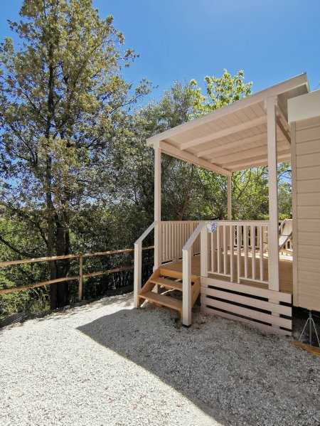 Veranda in legno affacciata su un giardino con alberi, sotto un cielo blu.