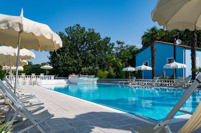 Piscina all'aperto con ombrelloni e lettini bianchi, circondata da alberi e un edificio blu.