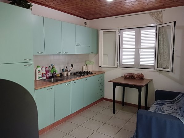 Cucina con mobili verde chiaro, finestre aperte e un piccolo tavolo con sedie.