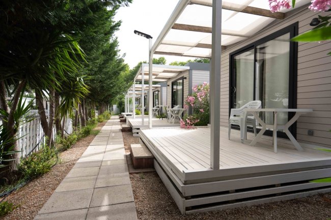 Area con bungalow moderni, terrazze in legno e giardino con alberi e fiori lungo un sentiero.