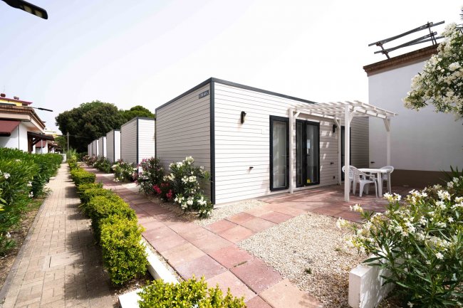 Bungalow moderno con terrazza, circondato da piante e fiori lungo un percorso pavimentato.