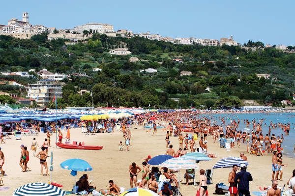 Una spiaggia affollata con ombrelloni colorati e un villaggio collinare sullo sfondo.
