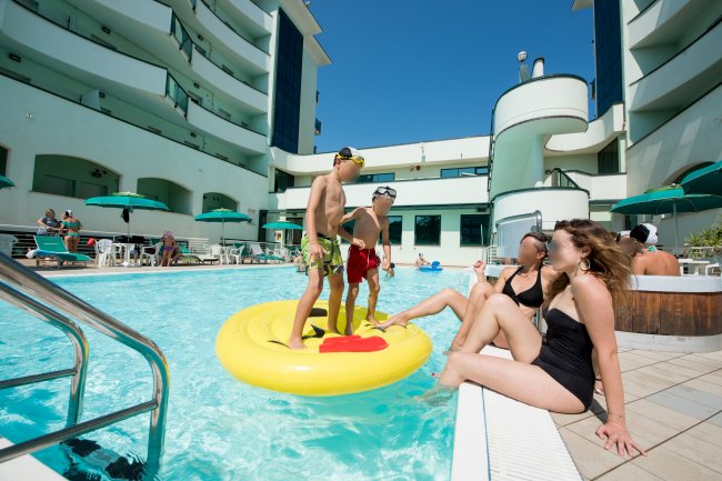 Due ragazzi su un materassino in piscina, due donne sul bordo, palazzi sullo sfondo.