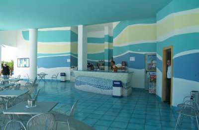 Spazio con tavolini e sedie, pavimento azzurro e pareti con strisce colorate, persone al bancone.