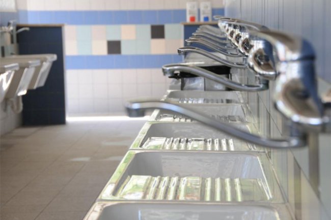 Fila di rubinetti sopra lavandini in un bagno pubblico con piastrelle a motivi chiari e scuri.