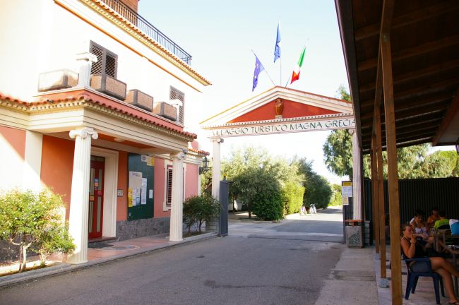 Ingresso con colonne bianche e bandiere, vegetazione circostante.