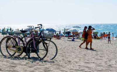 Biciclette parcheggiate sulla spiaggia, persone e ombrelloni sullo sfondo.