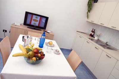 Cucina moderna con tavolo bianco imbandito e un cesto di frutta. Sullo sfondo, una TV accesa.