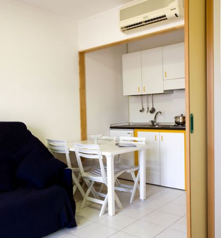 Piccola cucina con tavolo e quattro sedie pieghevoli bianche accanto a un divano scuro.