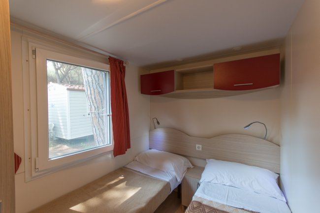 Interno di una casa mobile con due letti singoli, finestra e armadietti rossi.